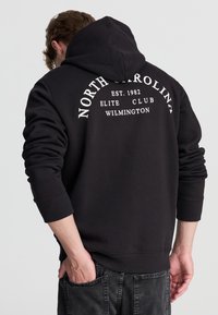 Czarna bluza z kapturem z dużym białym nadrukiem na plecach z napisem "NORTH CAROLINA EST. 1982 ELITE CLUB WILMINGTON", z prążkowanymi mankietami.