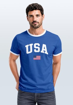 Mann mit dunklem lockigem Haar, der ein blaues T-Shirt mit weißem Rand und dem Text "USA" über einer kleinen amerikanischen Flagge trägt, steht mit den Händen in den Taschen.