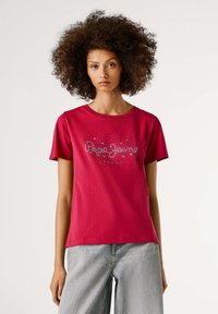 Rode katoenen T-shirt met korte mouwen, voorzien van een sprankelend tekstontwerp in zilver en donkergrijs, gecombineerd met lichtgrijze jeans.