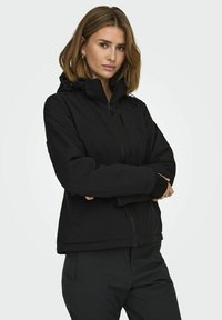 Veste à capuche noire avec une fermeture éclair à l'avant, en tissu lisse et mat. Elle possède des poches latérales et une coupe légèrement ample.