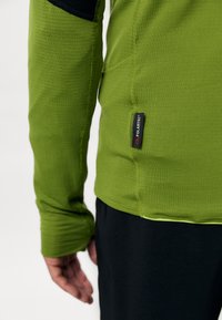 Zielona bluzka z długim rękawem, charakteryzująca się elastycznym dopasowaniem, teksturowanym materiałem oraz czarnym akcentem na ramieniu. Posiada metkę Polartec z boku.