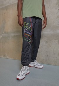 Man i svarta Nike trackpants med flerfärgat logo och vita sneakers med röda detaljer står mot en betongvägg.