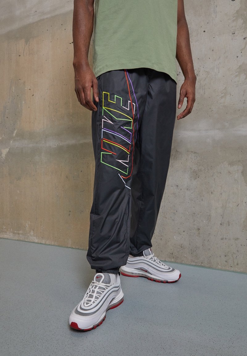 Man i svarta Nike trackpants med flerfärgat logo och vita sneakers med röda detaljer står mot en betongvägg.