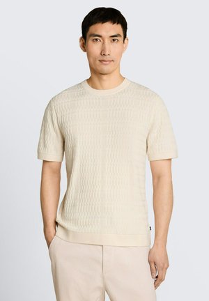 Mann trägt einen hellbeigen, strukturierten Kurzarmpullover und helle Hosen, steht mit einer Hand in der Tasche, neutraler Hintergrund.