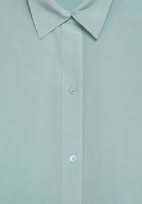 Chemise bleu clair à boutons avec col, présentant une texture lisse et une fermeture à un seul bouton. Design simple sans motifs.