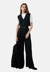 Zwarte cropped vest over een witte shirt, gecombineerd met hoge zwarte broeken en een riem. Model houdt een bijpassende zwarte jas vast.