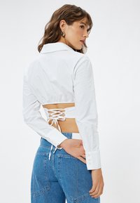 Hvid cropped skjorte med krave, lange ærmer og en samlet ryg med snøredetaljer. Kombinationen med blå denim jeans.