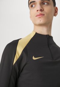 Fekete sportpóló arany kiegészítőkkel, magas gallérral, félig cipzárral és Nike logóval. Puha, rugalmas anyag, testhezálló fazon, sportolásra alkalmas.
