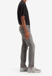 Jeans gris oscuro con un parche de cuero, combinados con una camiseta negra y zapatillas deportivas grises con acentos en blanco y rojo.