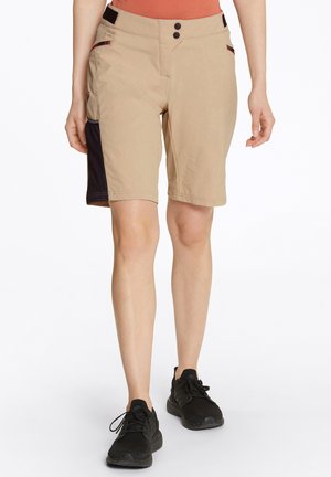 Beige shorts med en glat tekstur, to knapper foran, sidelommer og et mørkt stofpanel på venstre side. Sæt sammen med sorte sneakers.