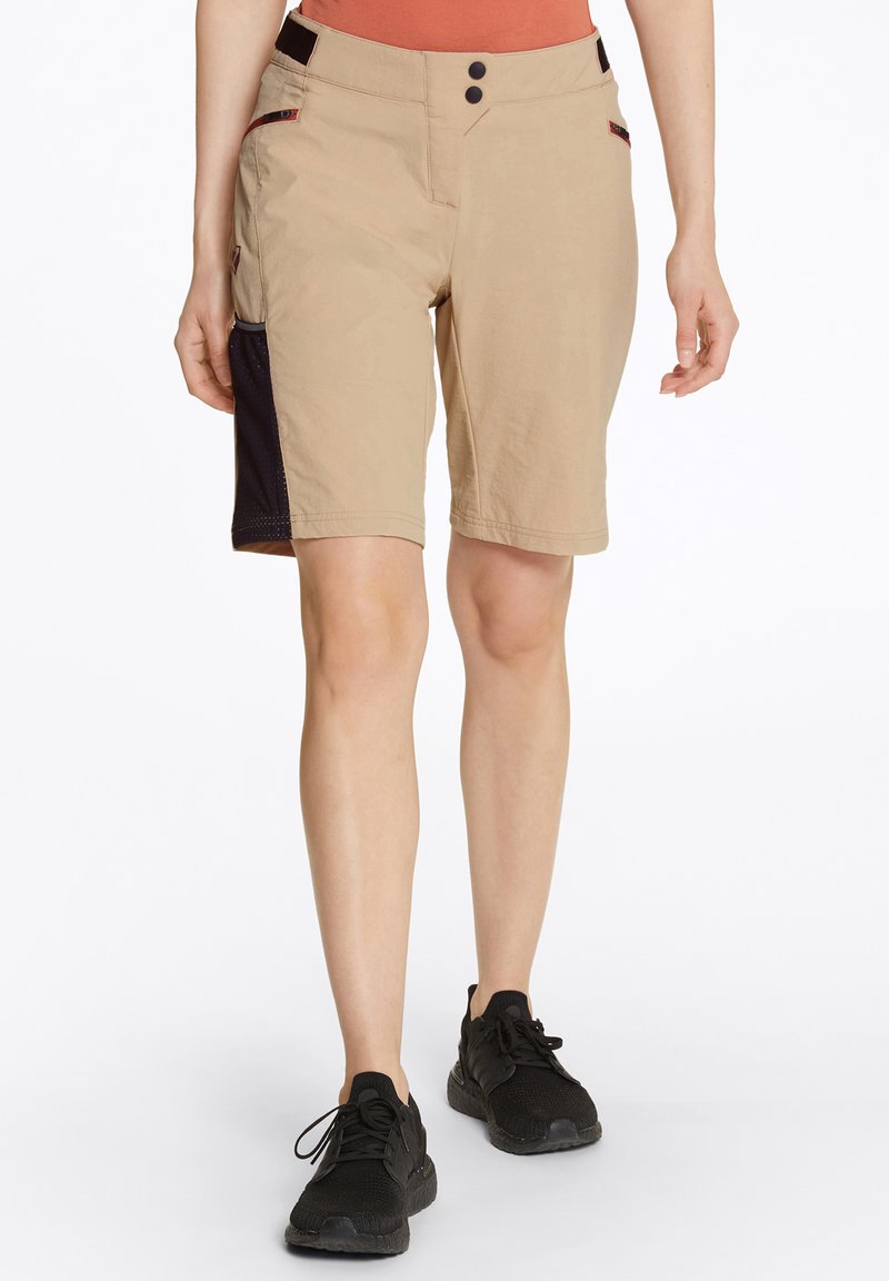 Beiga shorts med en slät yta, två knappar framtill, sidofickor och en mörk tygpanel på vänster sida. Matchas med svarta sneakers.