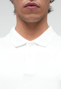 Cămașă polo albă cu guler, având trei butoane, țesătură texturată și un mic logo pe pieptul stâng.