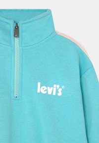 Sweatshirt em fleece azul claro com colarinho alto, meio zíper e um logotipo branco "Levi's" na frente. Apresenta detalhes em cores contrastantes nos ombros.