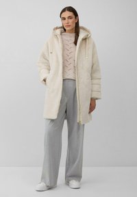 Crème-kleurig nepfleece jasje met een capuchon, voorzien van een rits en decoratieve trekkoorden, gecombineerd met een gebreide trui en grijze broek.