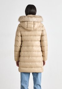 Person som bär en beige, vadderad pufferjacka med huva i pälskant, stående vänd bort mot en enfärgad vit bakgrund.