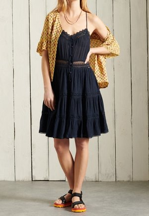 Femme portant une robe noire sans manches en dentelle jusqu'aux genoux, un châle jaune à motifs drapé sur les épaules, et des sandales noires avec des semelles orange.