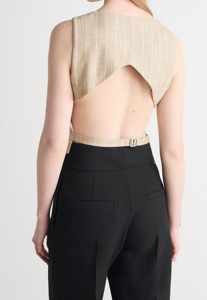 Vue de dos d'une personne portant un top court beige sans manches avec un dos ouvert et un pantalon noir taille haute avec un détail ceinture.