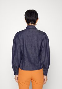 Camicetta in denim di colore blu scuro con maniche a sbuffo, polsini con bottone e colletto a punta. Texture liscia, vita aderente, indossata con pantaloni arancioni.