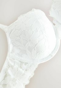 Reggiseno bianco in pizzo con motivo floreale, coppe imbottite e spalline regolabili. Presenta un intricato bordo in pizzo e dettagli in rete testurizzata.