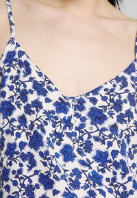 Närbild av en vit ärmlös topp med blå blommönster och tygklädda knappar i mitt fram.