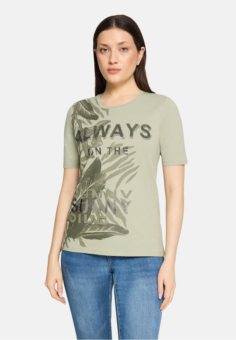 T-shirt en coton vert clair à manches courtes, avec un motif de feuilles et un texte en noir avec des accents métalliques violets.