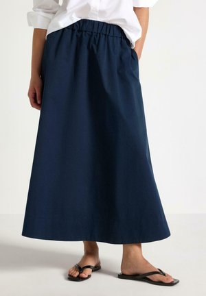 Donna che indossa una lunga gonna blu navy con vita elastica, abbinata a una camicia bianca e sandali infradito neri. Mani in tasca.