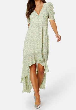 Femme portant une robe fleurie vert clair à coupe asymétrique, manches bouffantes, décolleté profond en V, boutons sur le devant et talons pointus argentés.