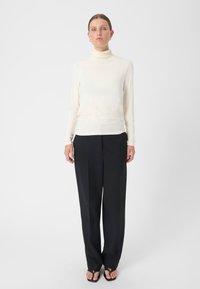 Langärmliger, cremefarbener Rollkragenpullover, kombiniert mit hoch-taillierten, weit geschnittenen schwarzen Hosen und schwarzen Flip-Flop-Sandalen. Einfaches, körperbetontes Design.