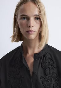 Close-up of a person with short blonde hair wearing a black embroidered shirt and a large gold hoop earring on the left ear.
