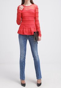 Top peplum rojo con mangas largas transparentes y textura de rayas horizontales, combinado con jeans ajustados azules y tacones negros. Cartera negra en mano.