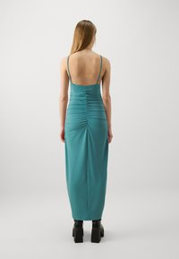 Teal ryggen fri kjole med spaghetti stropper, rynkede detaljer langs ryggen og en gulvlengde design. Svarte platå sko synlige.