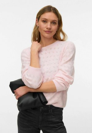 Femme aux cheveux blonds portant un pull en tricot rose clair et un jean noir, tenant une pochette en cuir noir.