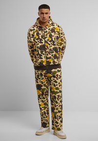 Giacca con cappuccio e zip e pantaloni coordinati in un motivo camouflage beige con accenti di faccine sorridenti gialle. Realizzati in un tessuto morbido, presentano una vestibilità rilassata.