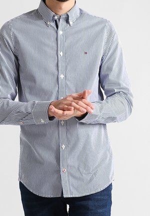 Homme portant une chemise à manches longues slim avec des rayures verticales noires et blanches, boutonnée, les mains jointes devant, jean foncé visible.