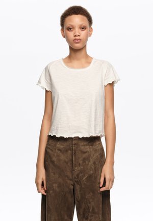 Joven con cabello corto que lleva una blusa blanca con volantes y mangas cortas, y pantalones marrones de textura, de cintura alta, de pie contra un fondo blanco.