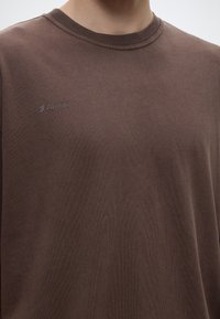 Καφέ t-shirt με στρογγυλή λαιμόκοψη και μικρό λογότυπο ARKK στο πάνω αριστερό μέρος του στήθους, που φοριέται από άτομο χωρίς εμφανές κεφάλι.