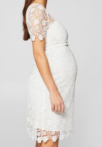 Vit spets gravidklänning med korta, mönstrade ärmar, blommönster, figursydd form och vågig nederkant. Inkluderar detalj med bältesmidja.