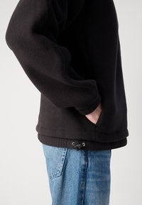 Pulover din fleece negru cu o suprafață texturată, având buzunare laterale și un șnur ajustabil la tiv, purtat cu blugi albaștri.