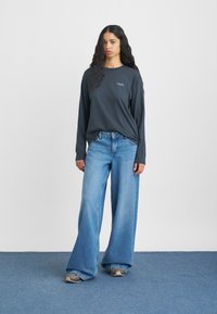 Czarna koszulka z długim rękawem z małym logo, zestawiona z szerokimi, jasnoniebieskimi dżinsami i szarymi trampkami na niebieskiej podłodze.