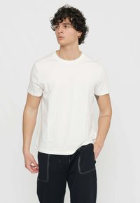 T-shirt en coton blanc avec un col rond et des manches courtes, présentant une coupe décontractée. L'arrière-plan est uni et neutre.