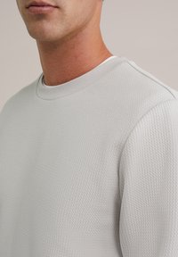 Sweatshirt gri deschis, texturat, cu guler rotund ribbat și detalii subtile de cusătură, prim-plan pe zona gâtului și a umărului.