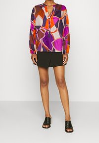 Blusa estampada de manga larga en tonos púrpura, naranja y rosa con patrones florales. Presenta un escote en forma de V y detalles de botones en la parte delantera.