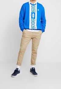 Sudadera azul con rayas blancas que lleva el logo "ADIDAS", combinada con pantalones chinos beige y zapatillas deportivas azul marino. Diseño sencillo, ajuste casual.