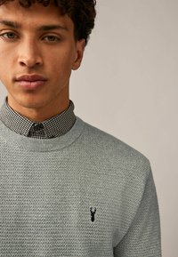 Pull en maille texturée bleu clair avec un col rond, arborant un emblème de cerf noir brodé sur la poitrine, superposé sur une chemise à carreaux noir et blanc.