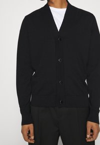 Svart stickad kofta med sjalkrage, femknäppning och ribbade ärmslut och nederkant. Tillverkad av mjukt, flexibelt material.