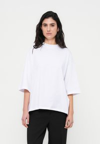 Άσπρο υπερμεγέθες μπλουζάκι με ψηλό λαιμόκοψη και κοντά μανίκια, κατασκευασμένο από απαλό, λείο ύφασμα. Συνδυάζεται με μαύρο παντελόνι.