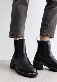 Czarne skórzane botki za kostkę z elastycznymi panelami bocznymi, słupkowym obcasem i białymi detalami szwów. W połączeniu z szarymi jeasami z podwiniętymi nogawkami i teksturowanymi skarpetkami.