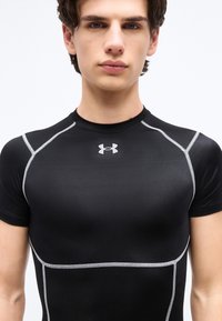 Νέος άνδρας που φοράει εφαρμοστό μαύρο αθλητικό πουκάμισο Under Armour με λευκή ραφή στους ώμους και στο στήθος, στραμμένος προς τα εμπρός.
