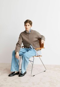 Bruine wollen jas met rits en twee voorzakken, gedragen over een blauw-wit gestreept overhemd, gecombineerd met lichtblauwe jeans en zwarte loafers.