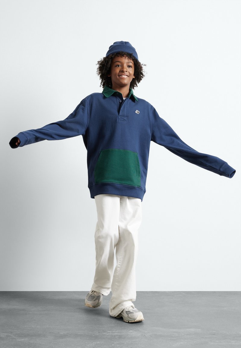 Glunderend kind in een blauw en groen sweatshirt, witte broek, grijze sneakers en een blauwe muts, met uitgestrekte armen wandelend over een grijze vloer.
